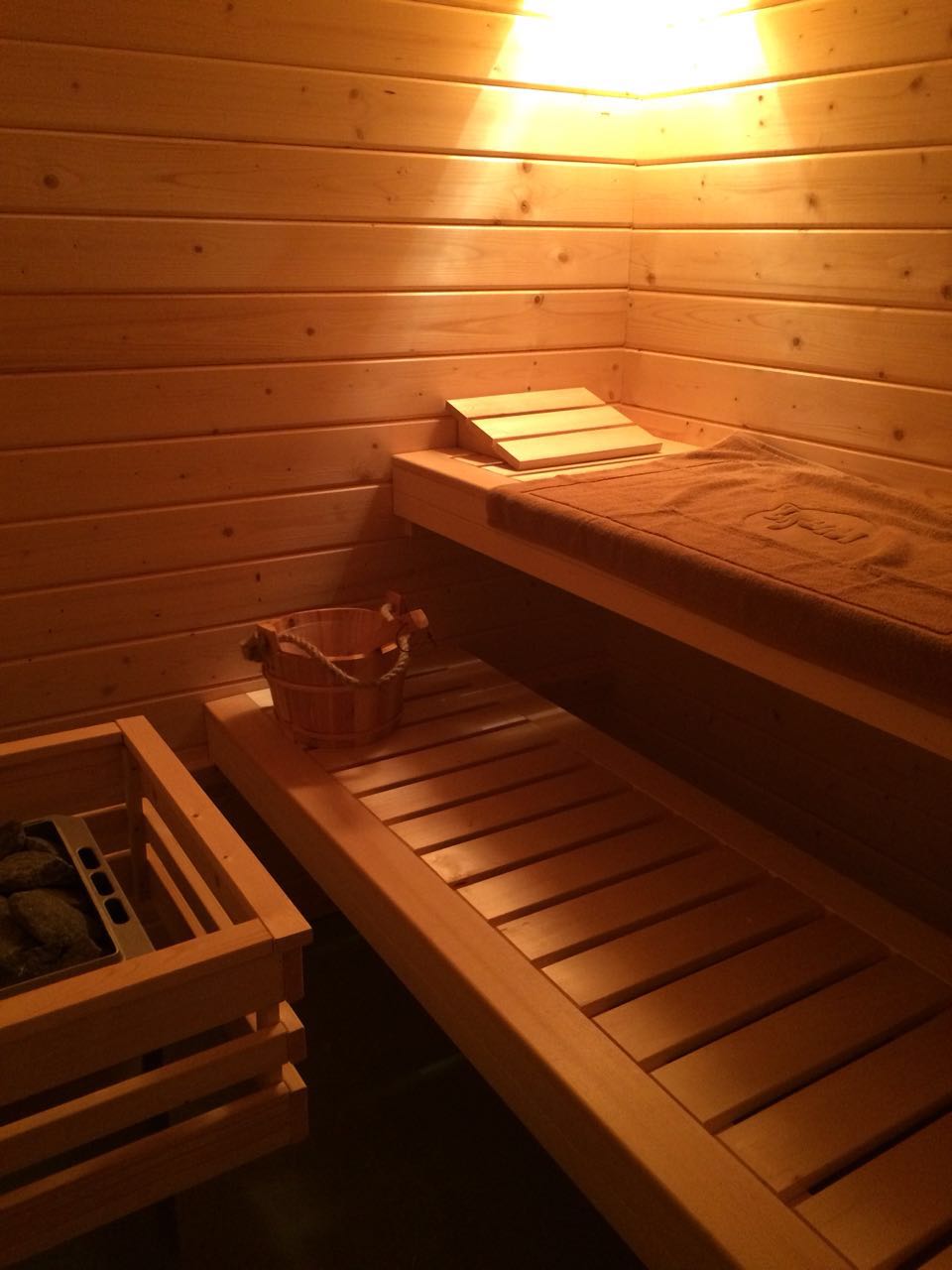 Innenansicht einer Sauna aus hellem Holz mit Bank, Aufgusskübel und Ofen