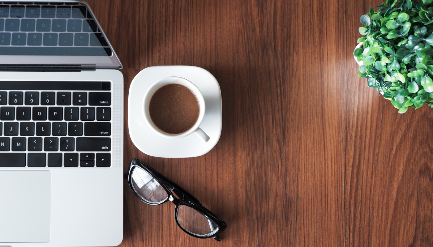 Laptop, Kaffeetasse und Brille auf einem warmen Holztisch