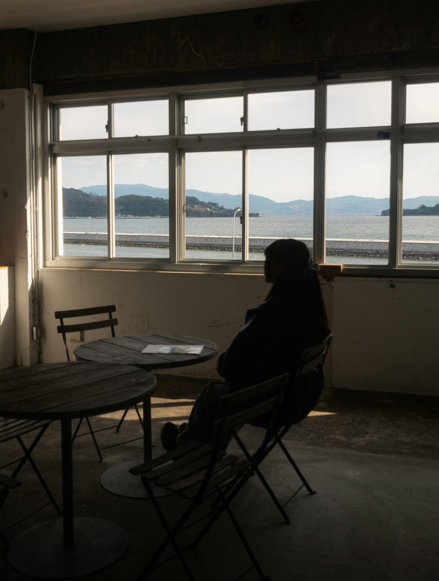Eine Person sitzt allein in einem dunklen Raum und schaut durch ein Fenster aufs Meer